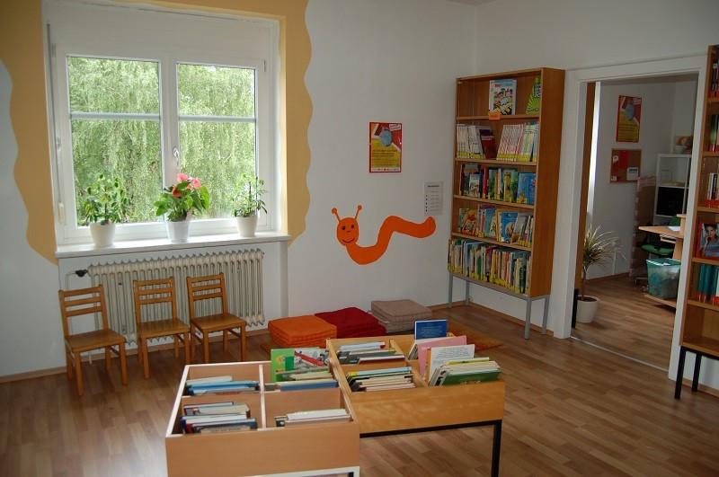 Kinderbereich mit Regal, 2 Kindersessel vor einem Fenster und Pölster an der Wand, Büchertröge davor